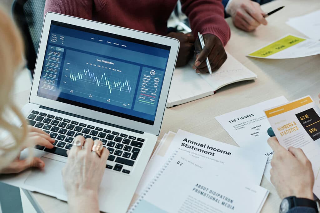 Hands working on a laptop displaying charts while documents and reports are spread out on the table.